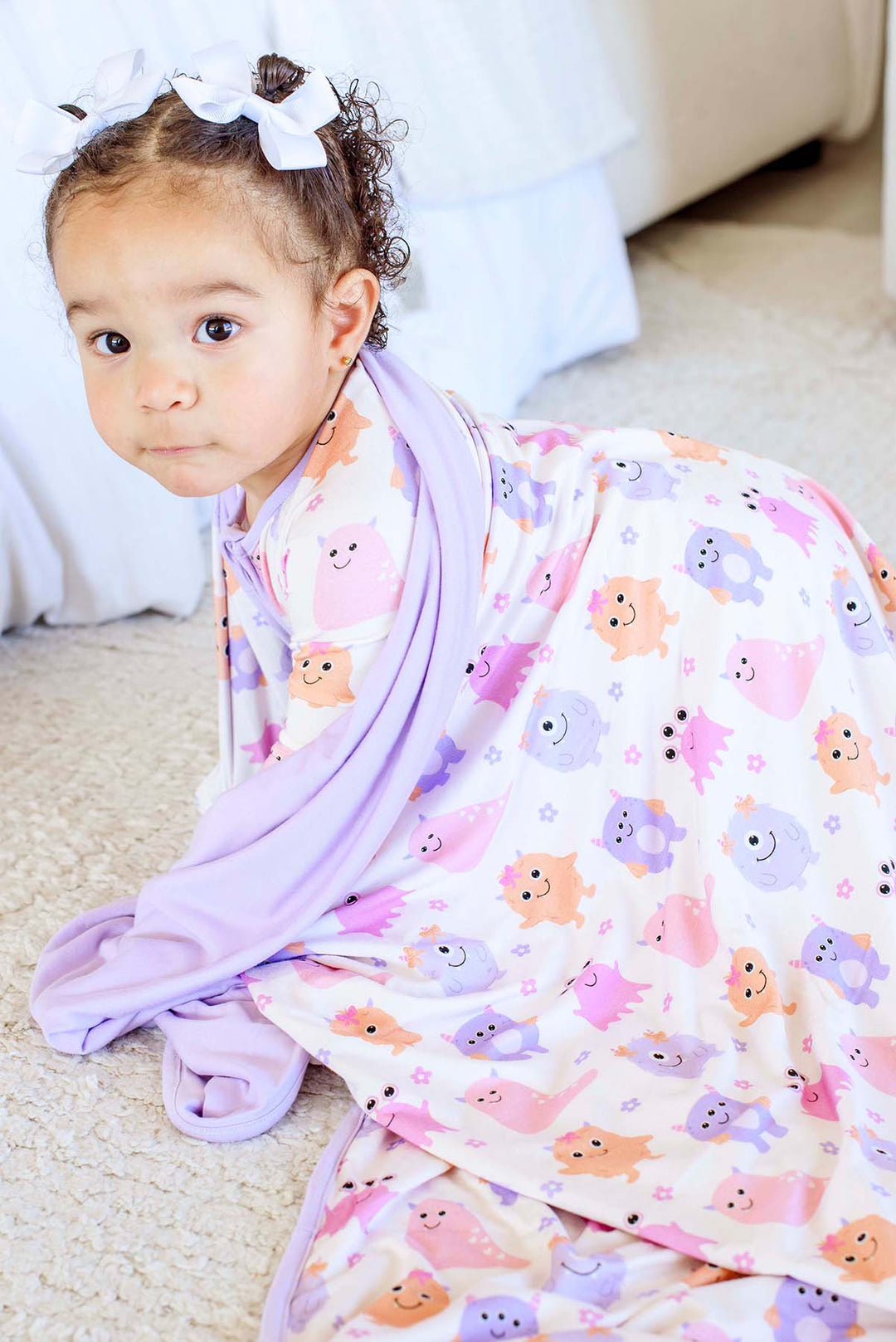 Toddler girl in white bows crawling while wrapped in a Caden Lane lavender-trimmed Little Monsters bamboo blanket, wearing matching monster-print pajamas