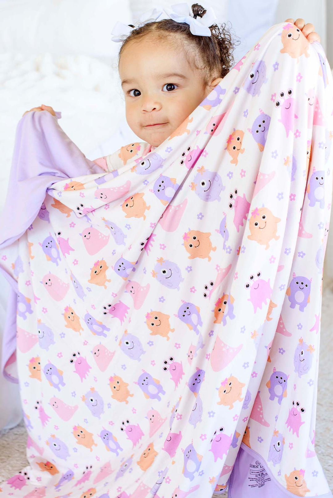 Toddler girl in a white bow holding up a Caden Lane lavender-trimmed Little Monsters bamboo blanket with pink, purple, and orange cartoon monsters