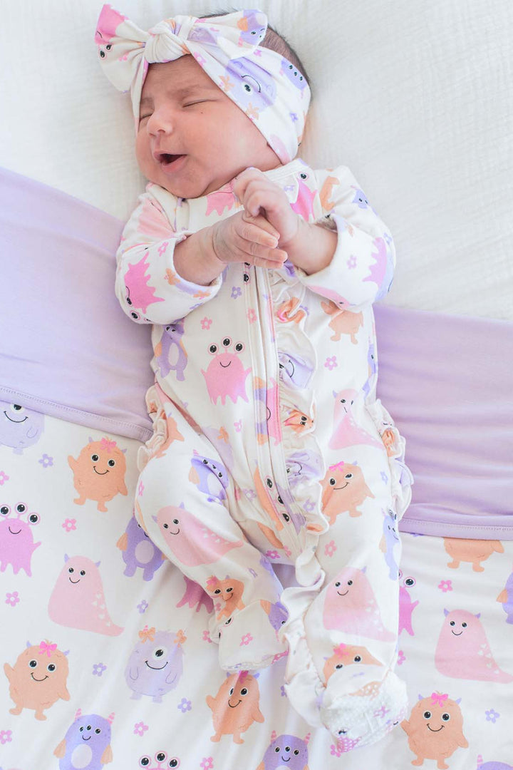 A sleeping newborn baby wearing a white footed sleeper and matching headband with a pink, purple, and orange monster pattern, lying on a white and lavender mattress.