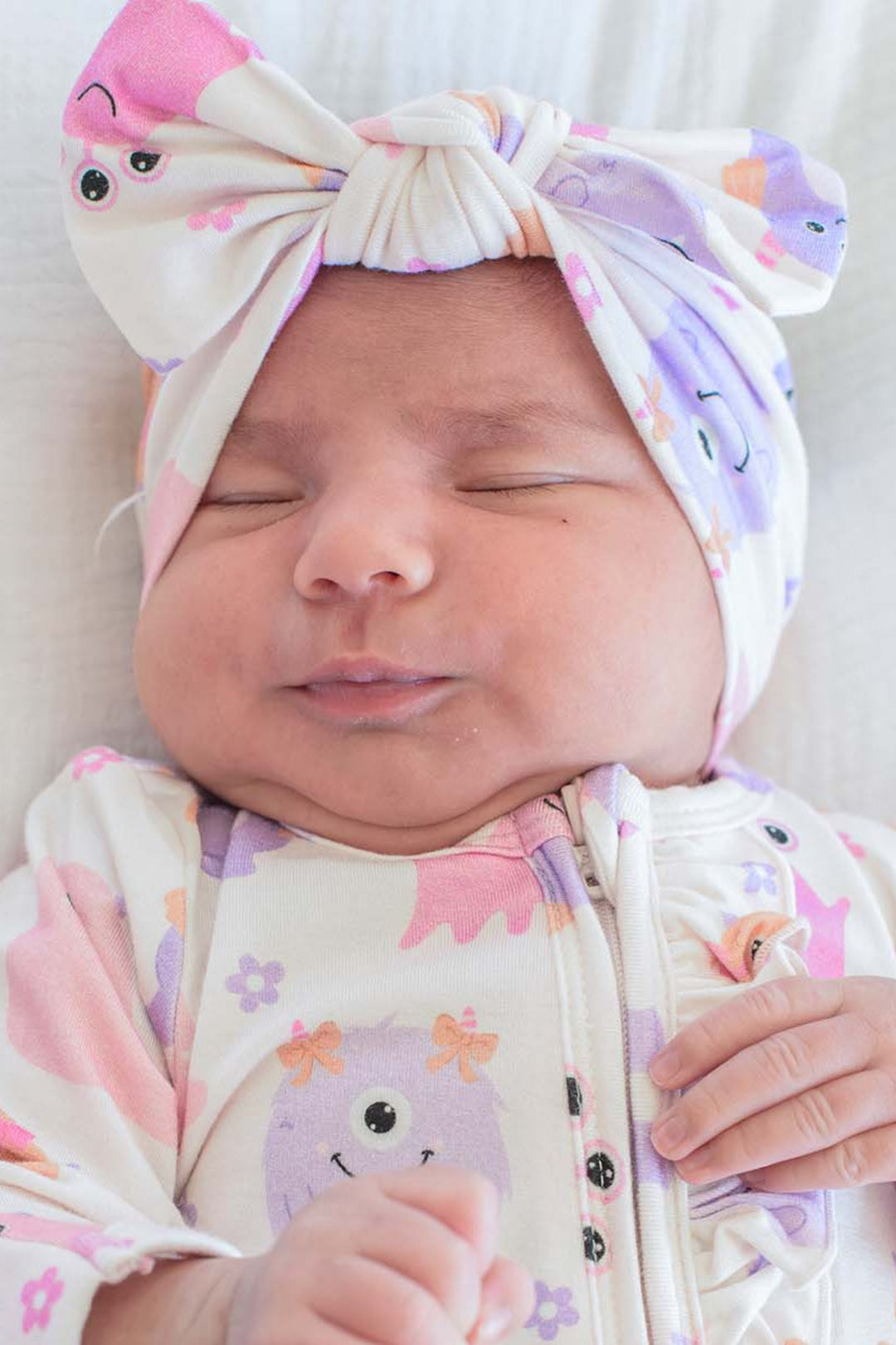 A sleeping newborn baby wearing a cream-colored zip-up footed sleeper with a pink, purple, and orange cartoon monster print, along with a matching knotted turban hat.