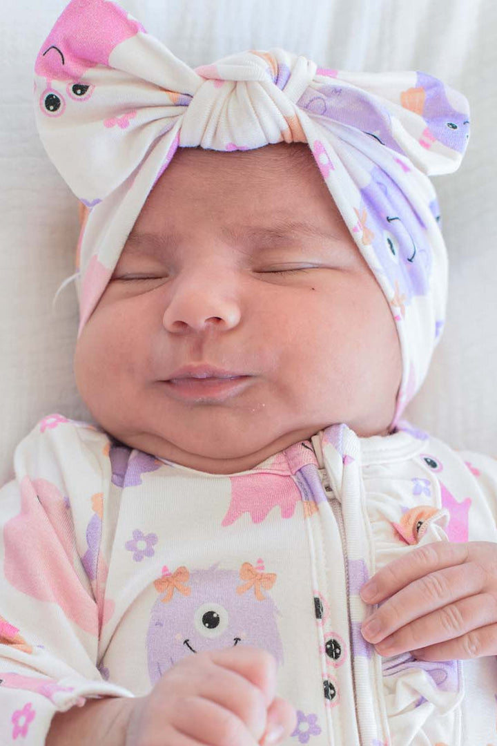 A sleeping newborn baby wearing a cream-colored zip-up footed sleeper with a pink, purple, and orange cartoon monster print, along with a matching knotted turban hat.