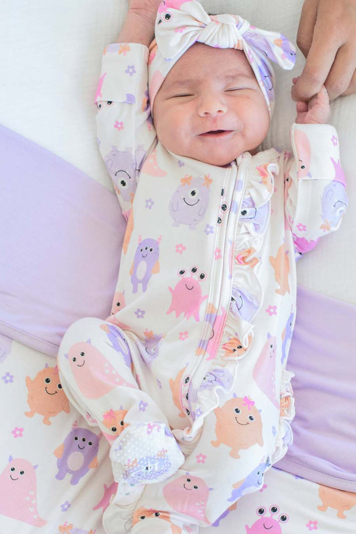 Smiling newborn held by parents' hands, wearing a Caden Lane Little Monsters ruffle footie with matching bow headwrap