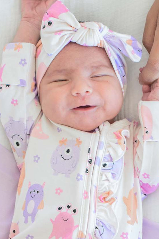 A sleeping newborn baby wearing a cream-colored footed onesie and matching headband with a pink, purple, and orange monster pattern, lying on a white and lavender changing pad while being held by an adult's hand.