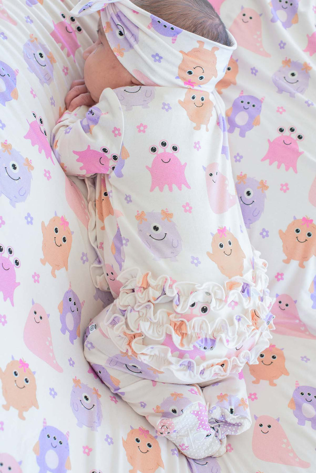 A baby wearing a Caden Lane white footed romper with a ruffled hem and matching head wrap, lying on a bed with a matching monster-print bedding set. The print features cute pink, purple, and orange cartoon monsters on a white background.