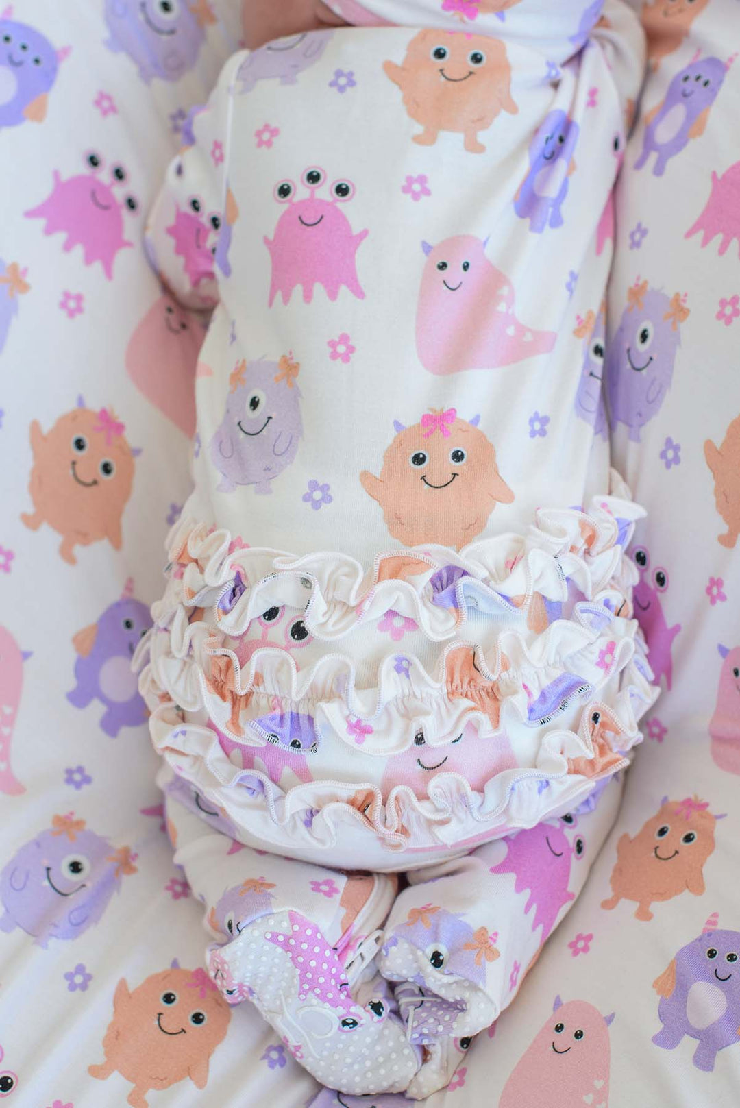 A baby swaddled in a soft cream fabric with a playful print of smiling cartoon monsters in pink, purple, and peach, featuring a ruffled trim around the waist and matching foot covers.