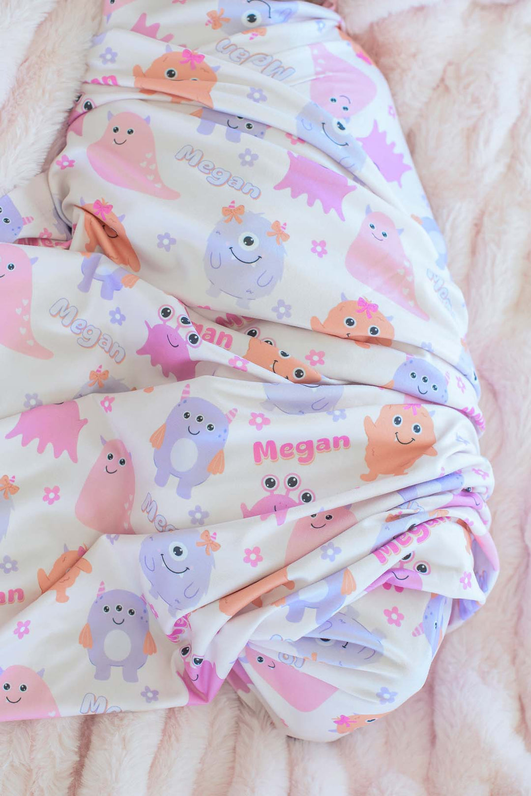 Close-up of Caden Lane personalized "Megan" swaddle blanket in Little Monsters print with pink, purple, and orange cartoon monsters and flowers, folded on a pink faux fur blanket