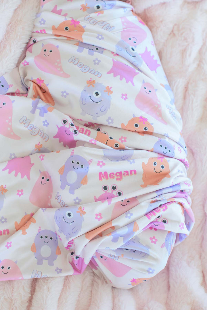 Close-up of Caden Lane personalized "Megan" swaddle blanket in Little Monsters print with pink, purple, and orange cartoon monsters and flowers, folded on a pink faux fur blanket