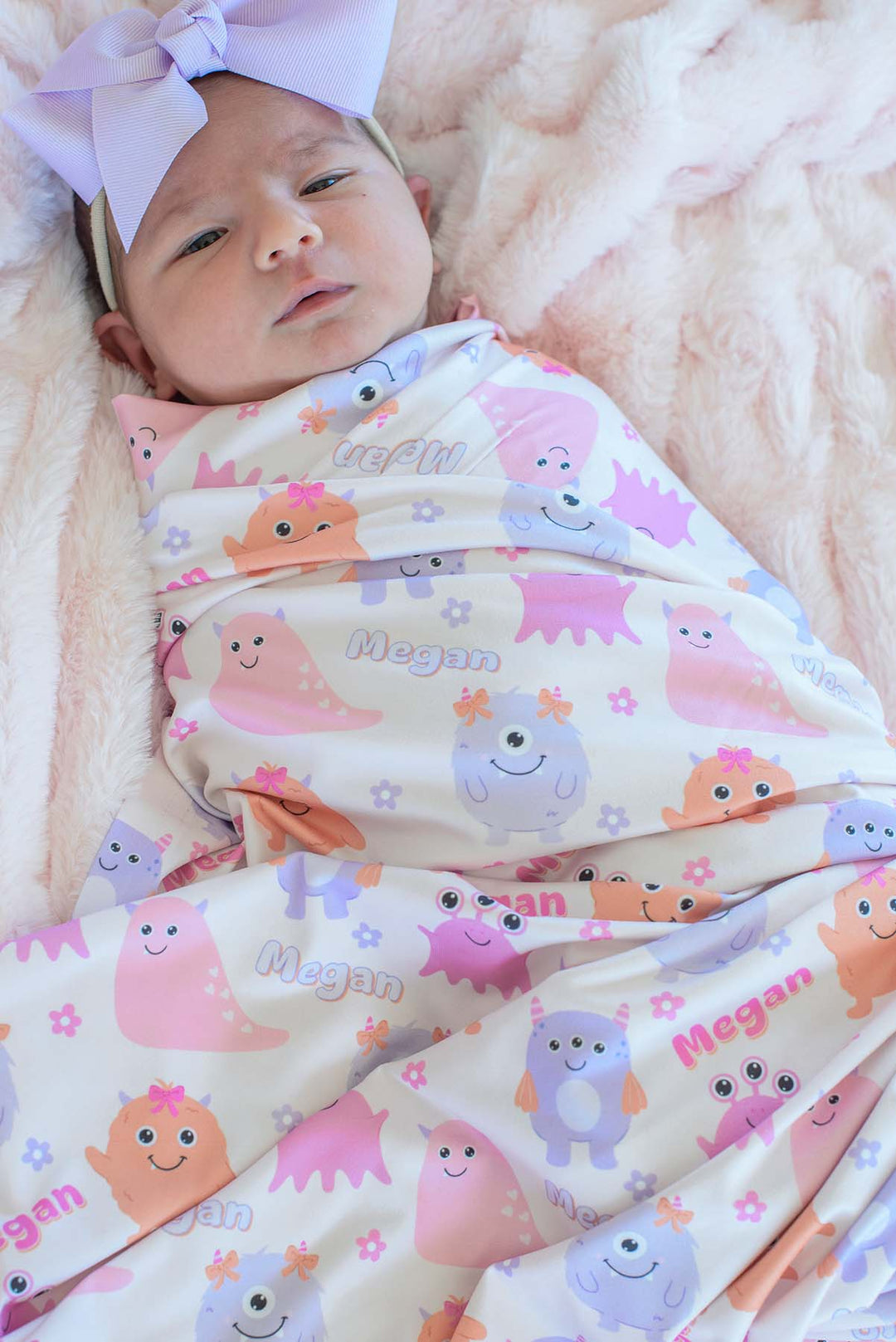 Baby wearing a lavender bow headband swaddled in Caden Lane personalized "Megan" Little Monsters print blanket with pink, purple, and orange cartoon monsters