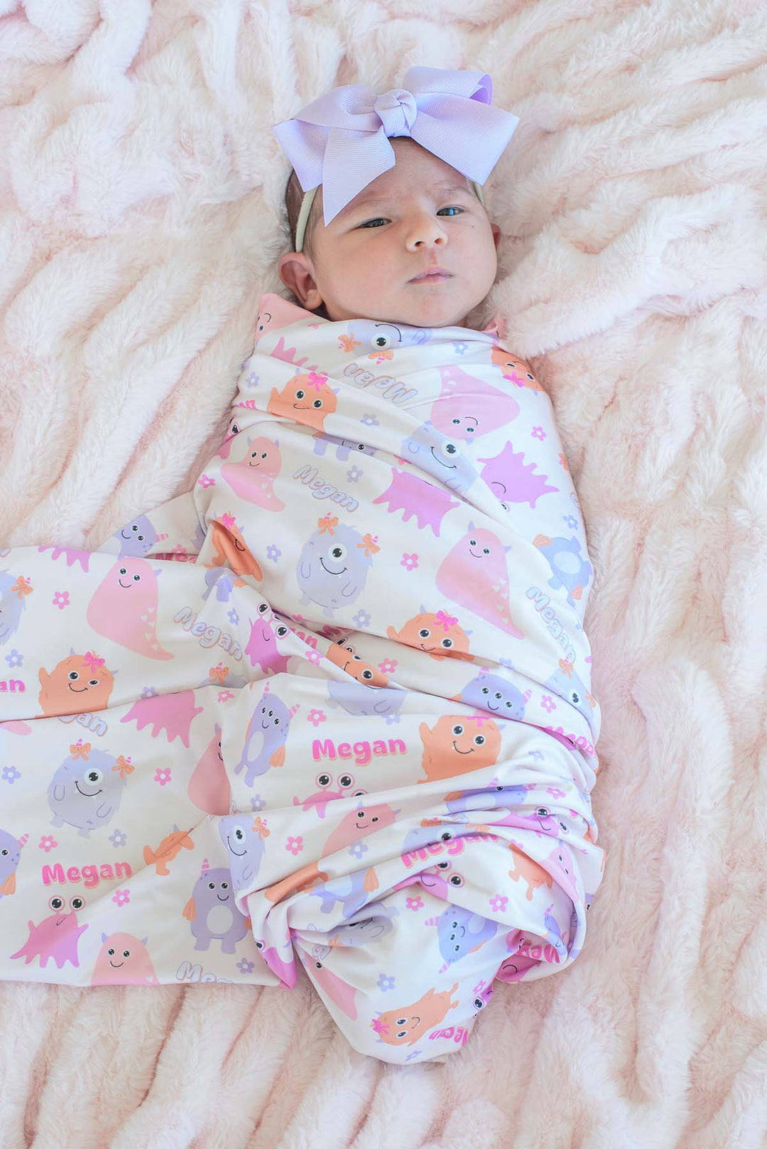 Baby wearing a lavender bow headband swaddled in Caden Lane personalized "Megan" Little Monsters print blanket, overhead view on a pink faux fur backdrop