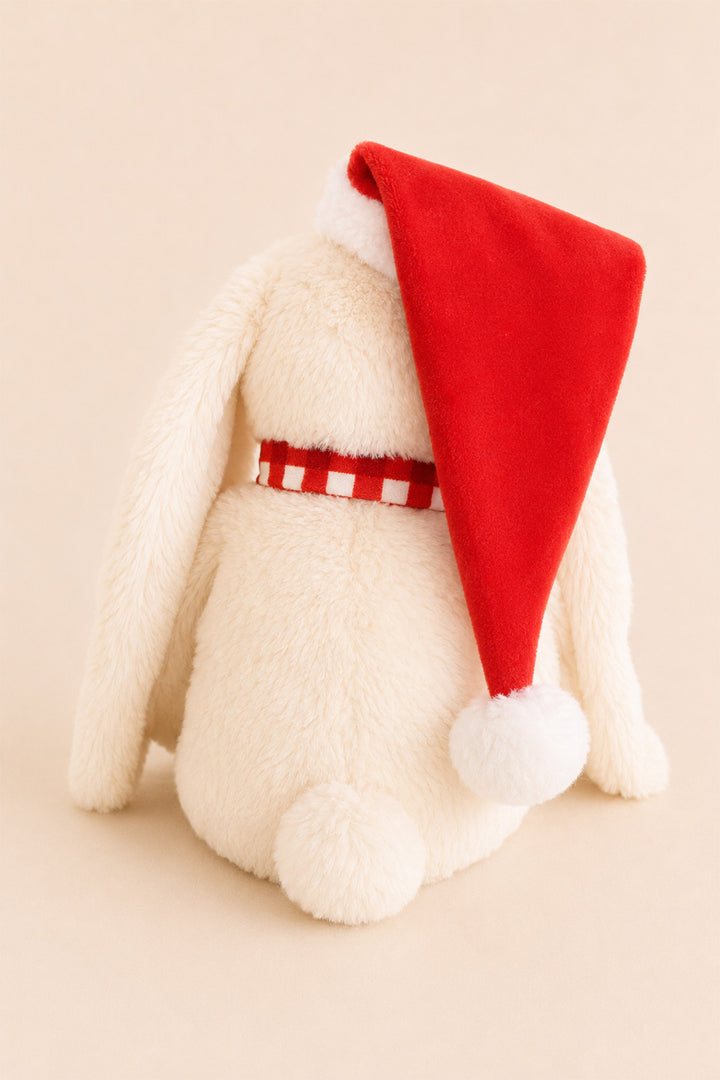 A Caden Lane cream-colored plush bunny toy wearing a red Santa hat with a white pom-pom and a red-and-white checkered belt, photographed against a warm beige background.