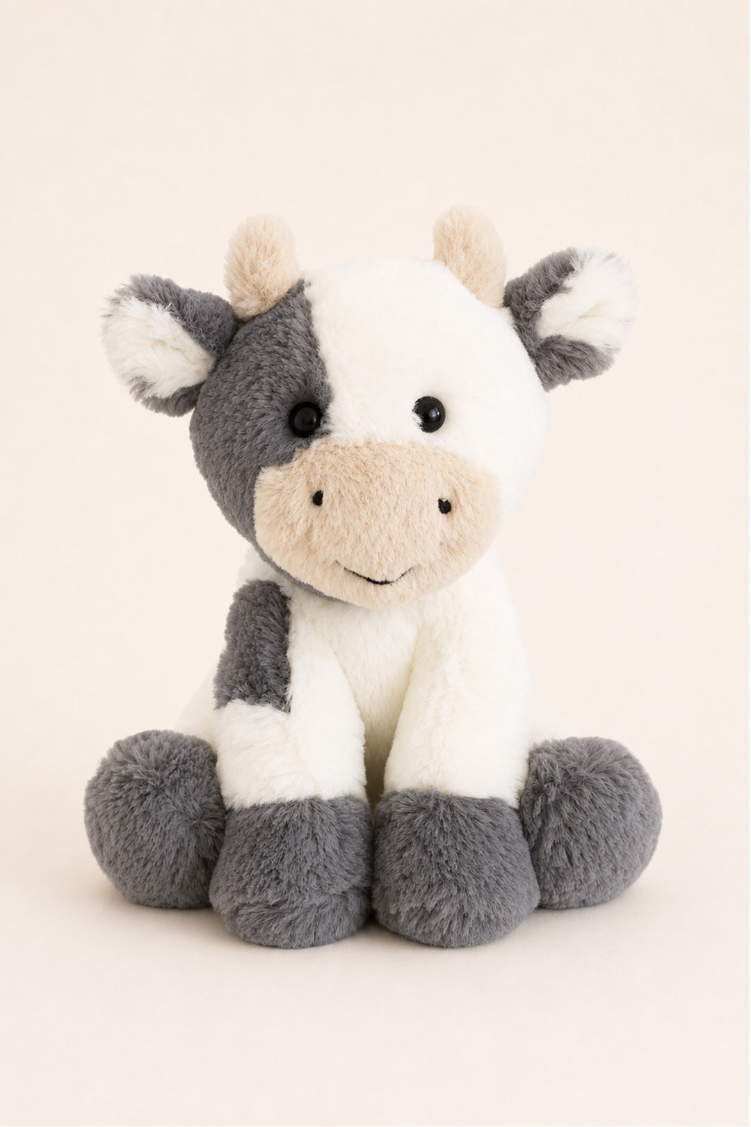 A Caden Lane plush cow toy with a white and gray color-blocked pattern, featuring a beige snout and ears, sitting against a solid light beige background.