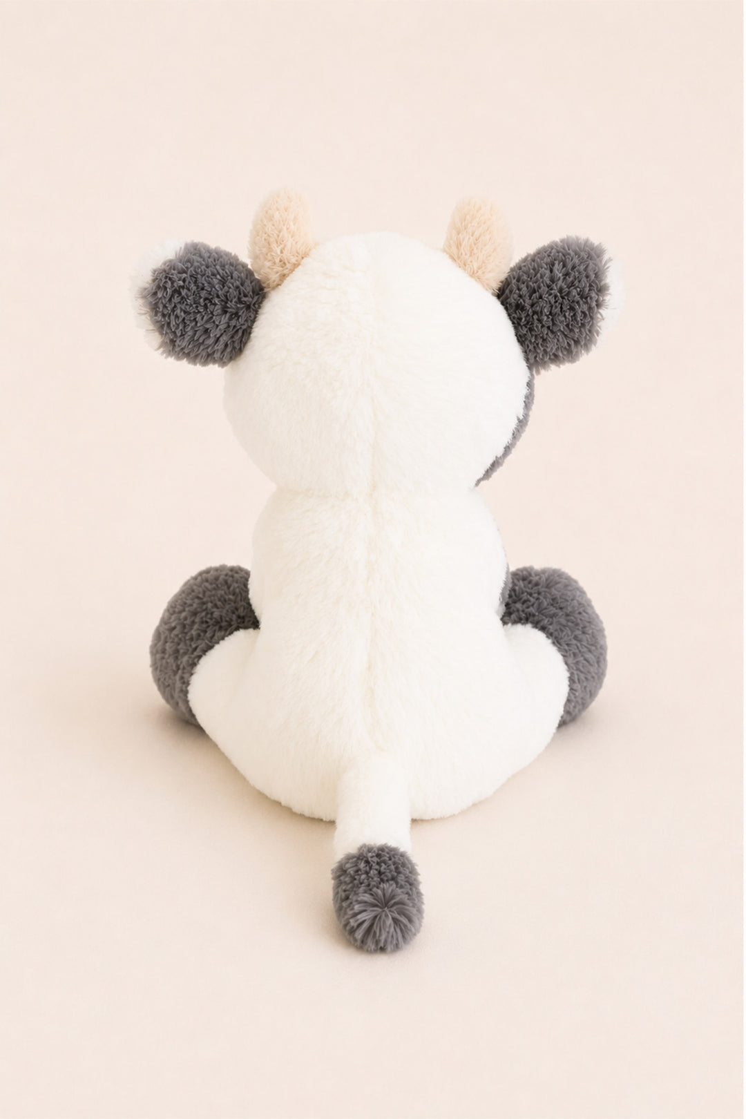A Caden Lane plush cow toy shown from the back, featuring a white body, dark gray ears, hooves, and tail, and light beige horns, set against a solid pale pink background.