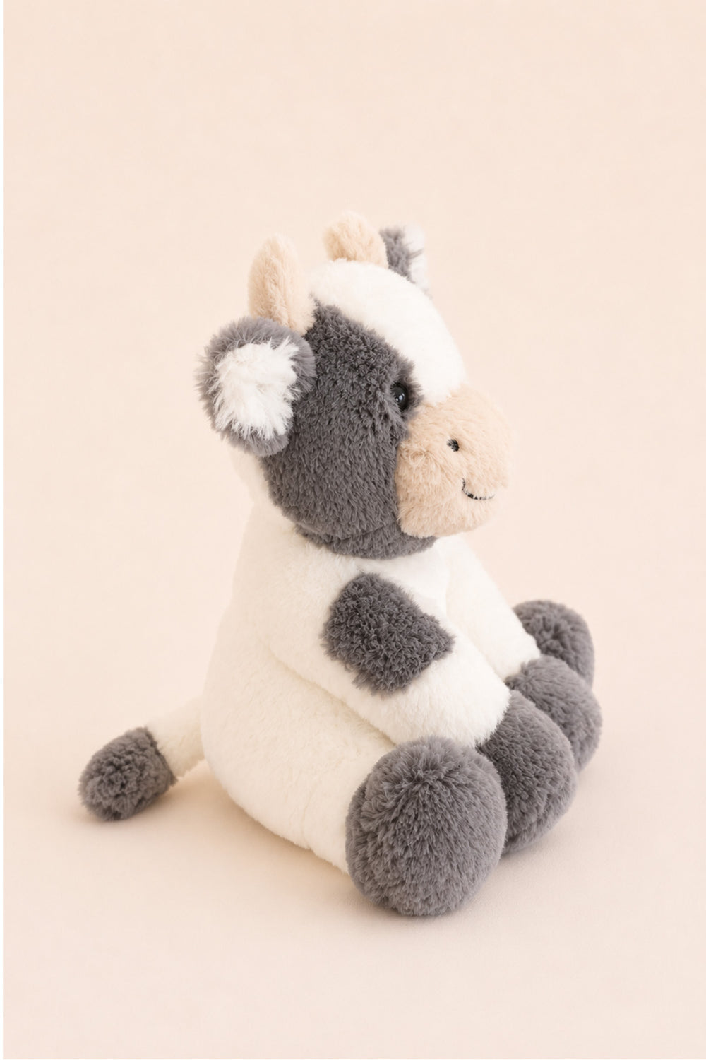 A Caden Lane plush cow toy with a white body, gray patches, and a beige snout, sitting against a solid pale peach background.