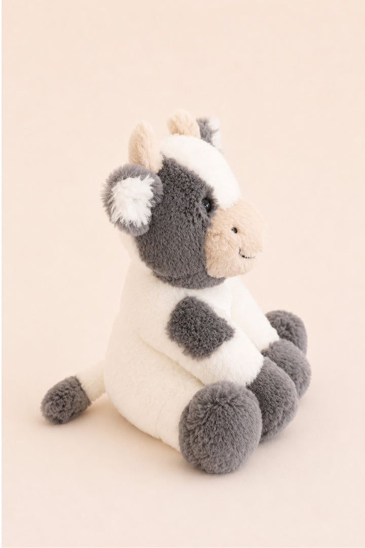 A Caden Lane plush cow toy with a white body, gray patches, and a beige snout, sitting against a solid pale peach background.