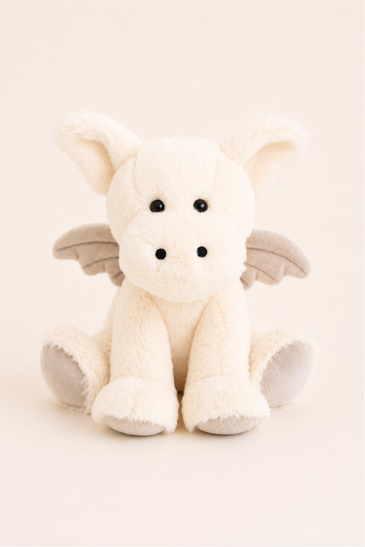 A Caden Lane plush dragon toy in off-white faux fur with small gray wings and black bead eyes, sitting against a solid cream background.
