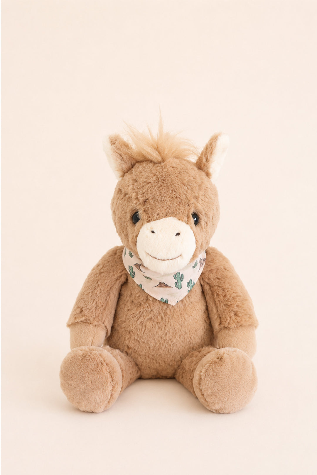 A Caden Lane plush toy horse sitting upright against a solid, warm off-white background. The toy is made of soft brown plush fabric with a white muzzle, embroidered black eyes and nose, and a patterned bandana around its neck featuring green cactus motifs.