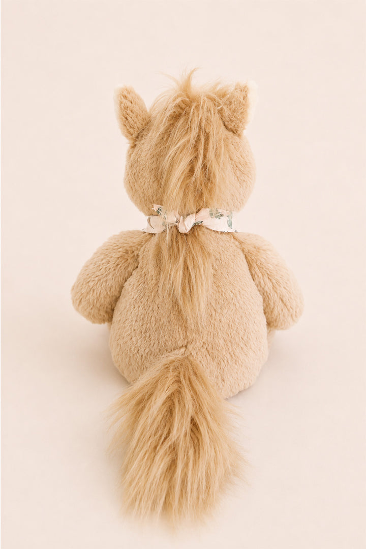A Caden Lane plush stuffed animal shaped like a horse or pony, shown from the back on a plain warm beige background. The toy is light tan/beige with a small white bow around its neck.
