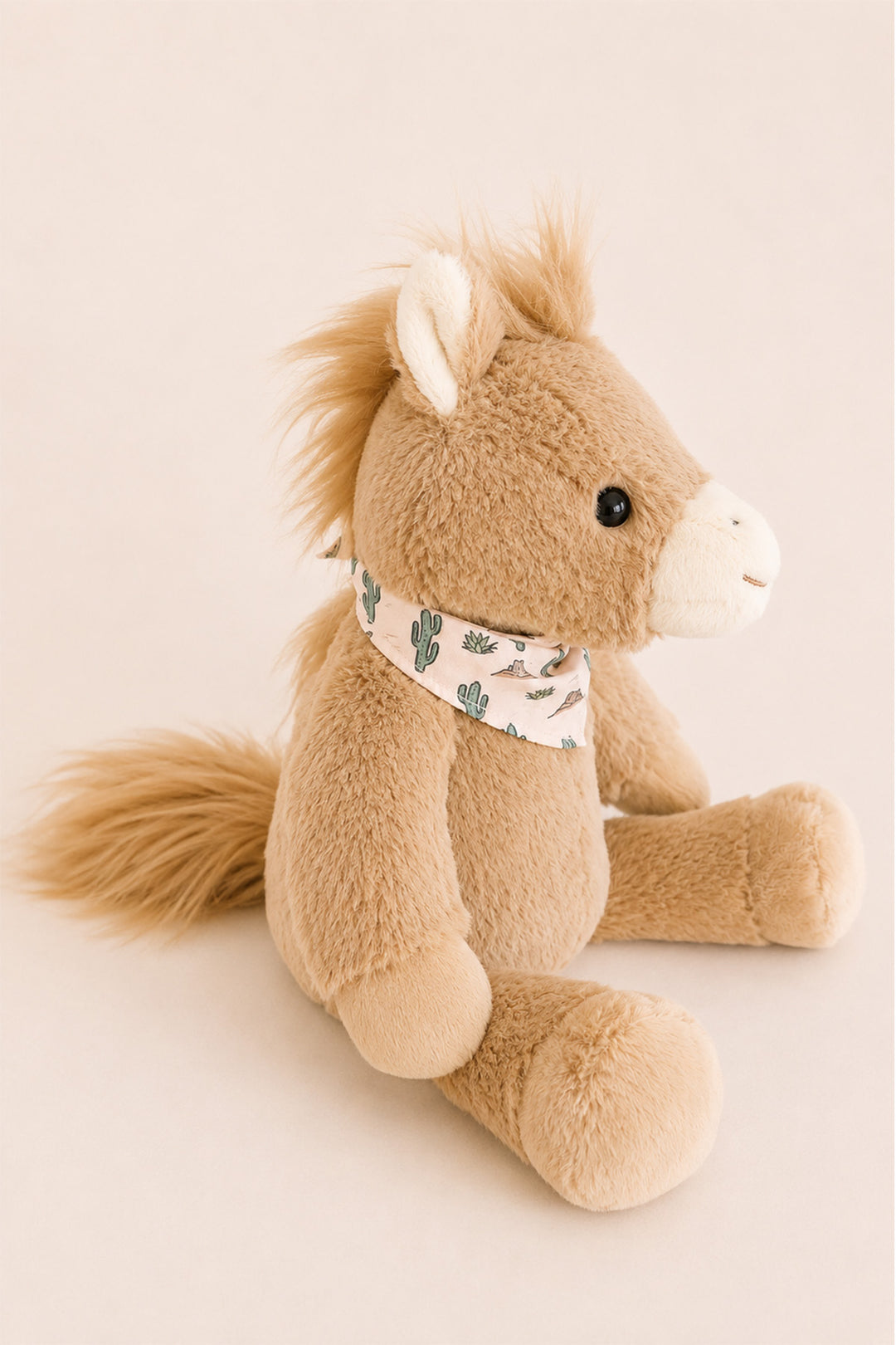 A Caden Lane plush toy horse in light tan with a white snout, black eyes, and a cream bandana patterned with small green cacti, sitting against a solid pale beige background.