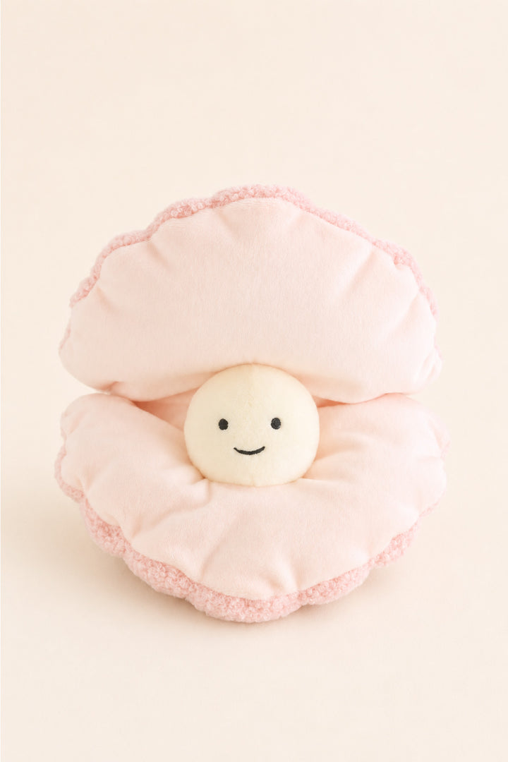 A Caden Lane plush pillow shaped like an open pink clam with a small smiling cream pearl character in the center, photographed on a soft cream background.