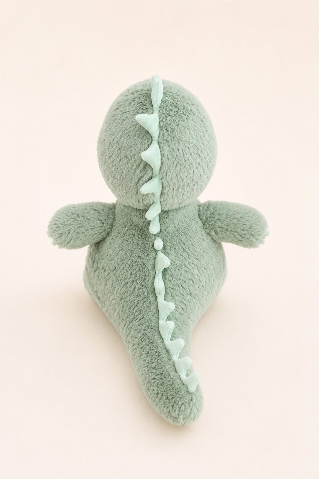 A mint green Caden Lane plush dinosaur toy shown from the back, featuring a textured fuzzy body and a lighter mint green spiky back detail, set against a solid pale pink background.