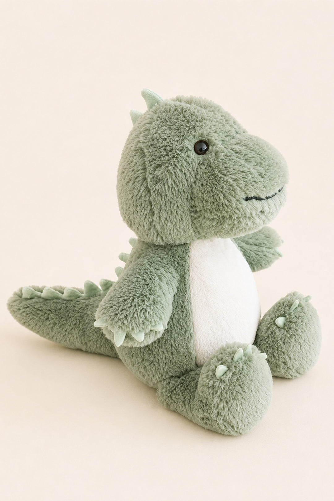 A plush green Caden Lane dinosaur toy with a white belly, sitting on a plain beige background.