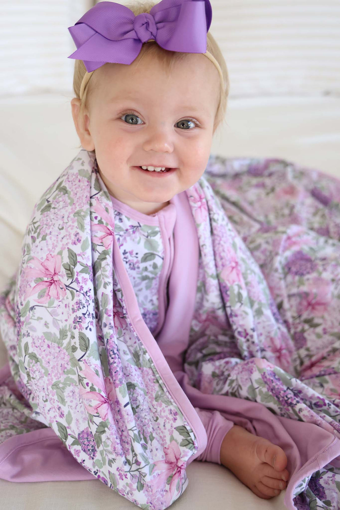 sitting baby girl with lavender lullaby bamboo blanket around her shoulders wearing matching pajamas and a purple ribbon bow headband