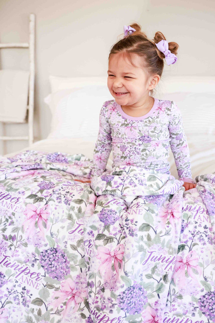 smiling girl sitting on bed in lavender lullaby two piece pajama set with personalized blanket across her lap