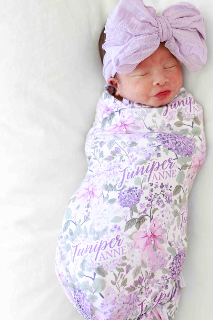 newborn girl wrapped in lavender lullaby personalized swaddle blanket with purple ruffle bow on her head