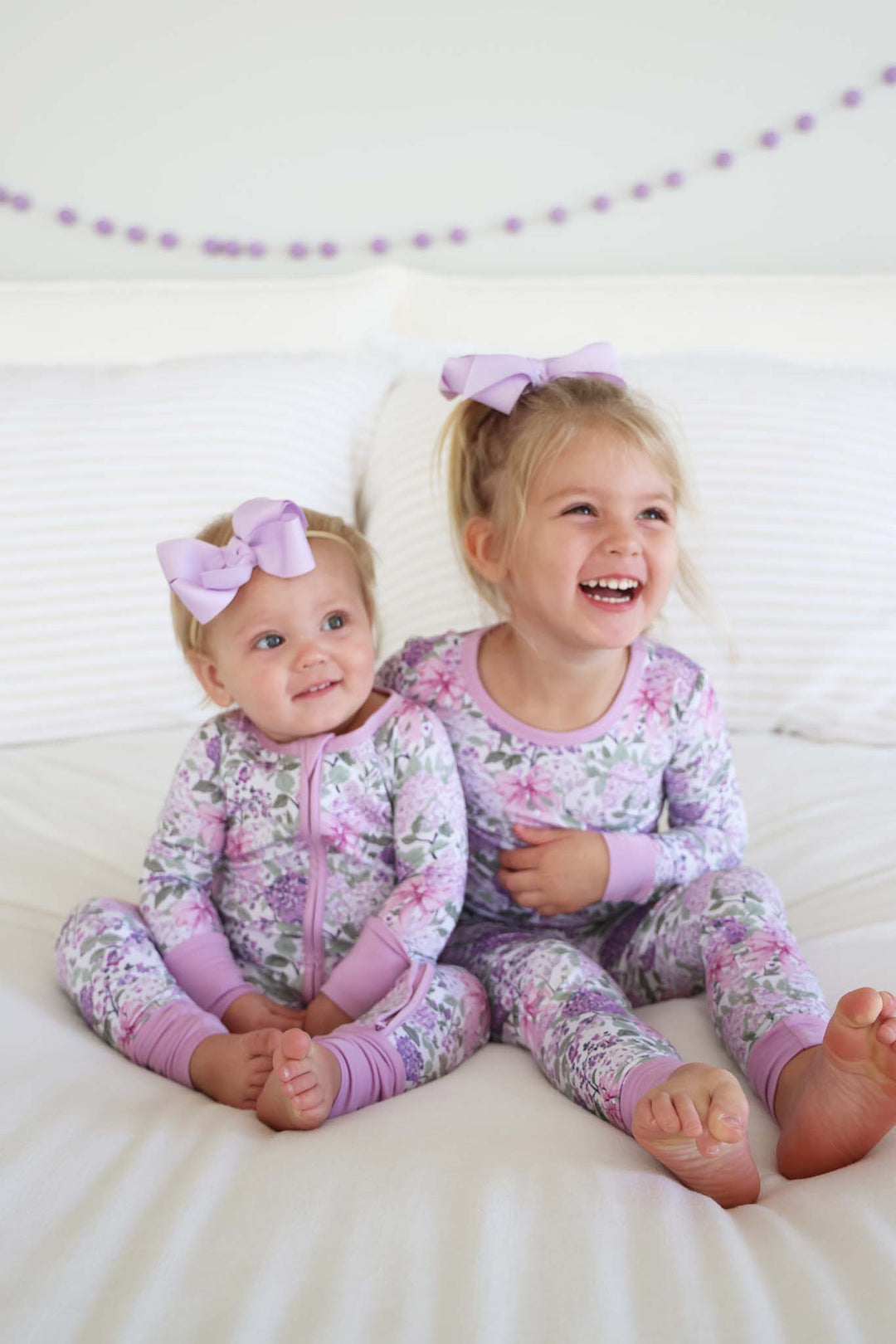 sisters sitting on bed in lavender lullaby convertible zip romper and two piece pajama set