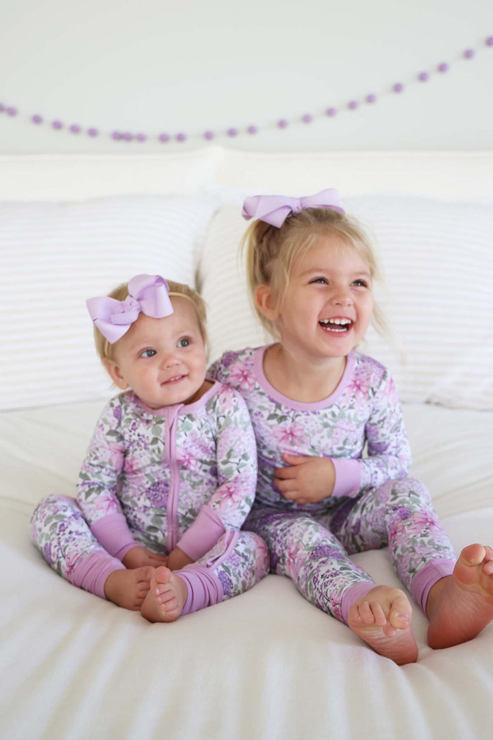 sisters sitting on bed in lavender lullaby convertible zip romper and two piece pajama set