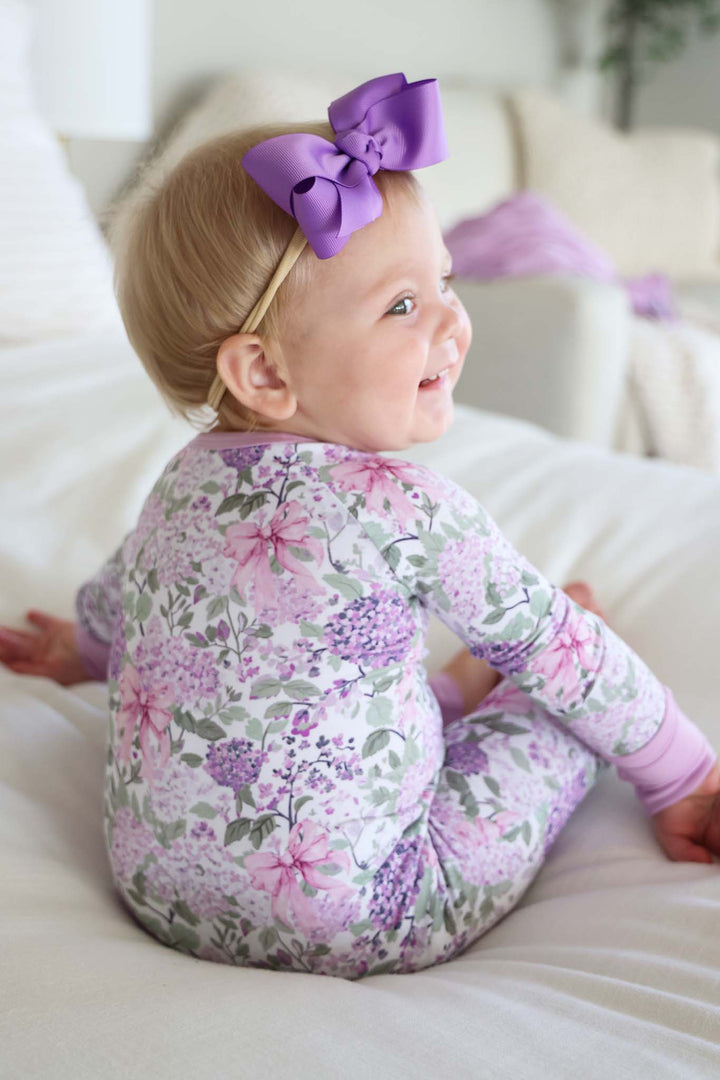 baby girl looking over her shoulder in purple hydrangea and pink bow pajama romper and amethyst ribbon bow headband
