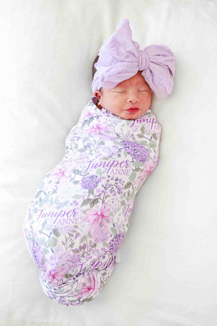 baby girl laying on pillow in lavender lullaby personalized swaddle with matching purple ruffle knit bow