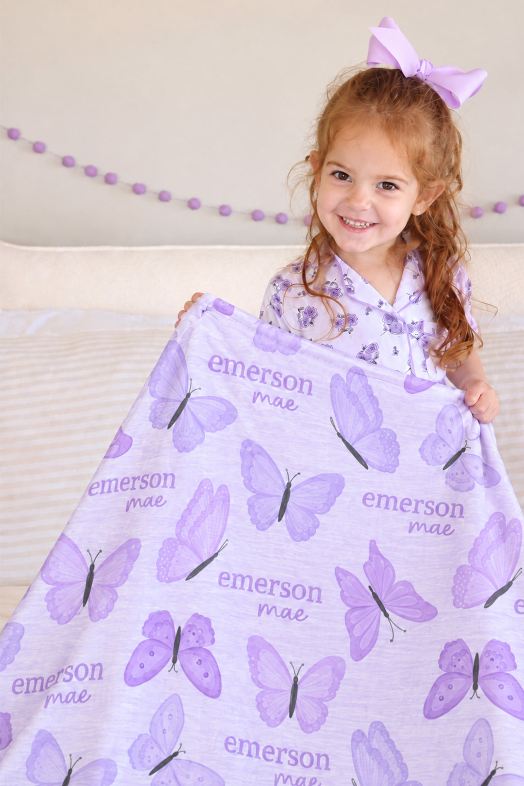 Girl in floral pajamas holding up a personalized Caden Lane Lavender Wings blanket, showing purple butterflies and custom name print