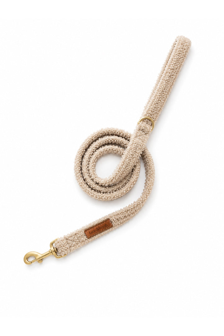 A Lane & Leash  beige bouclé rope dog leash with a gold-tone metal swivel snap hook and a small brown leather brand patch, shown coiled on a white background.