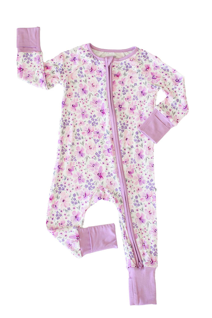 Flat lay of a Caden Lane white knit zip romper with fold-over lilac cuffs in a Lilac Dreams print featuring purple watercolor flowers and grey leaves.