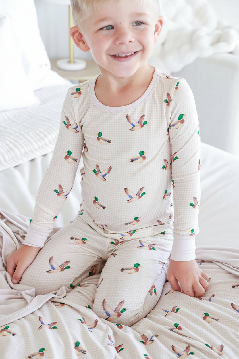 Smiling toddler kneeling on a bed wearing a Caden Lane cream bamboo two-piece pajama set in a Little Mallard print featuring flying mallards on gingham, paired with a matching blanket.