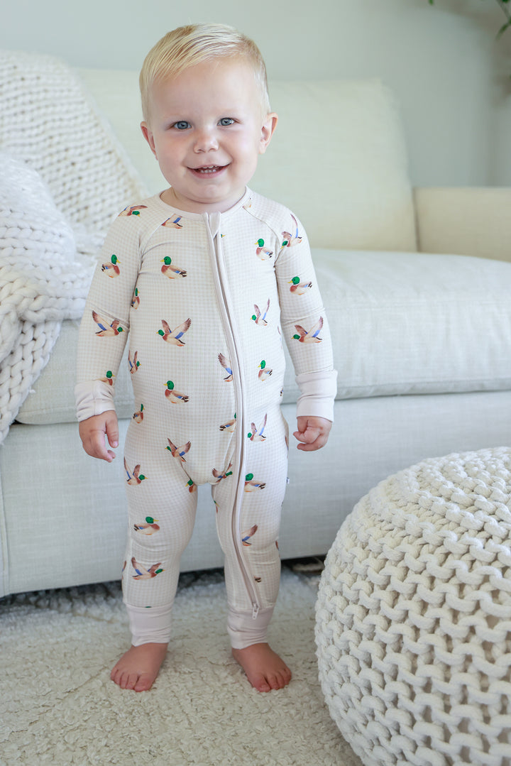 Toddler standing and smiling wearing little mallard duck print convertible zip romper, front view showing full-length zip and duck print