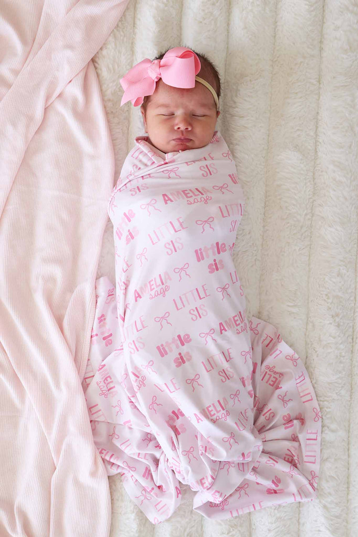 baby girl laying on white fluffy blanket wrapped in little sis bow personalized swaddle blanket wearing a pink ribbon bow headband 