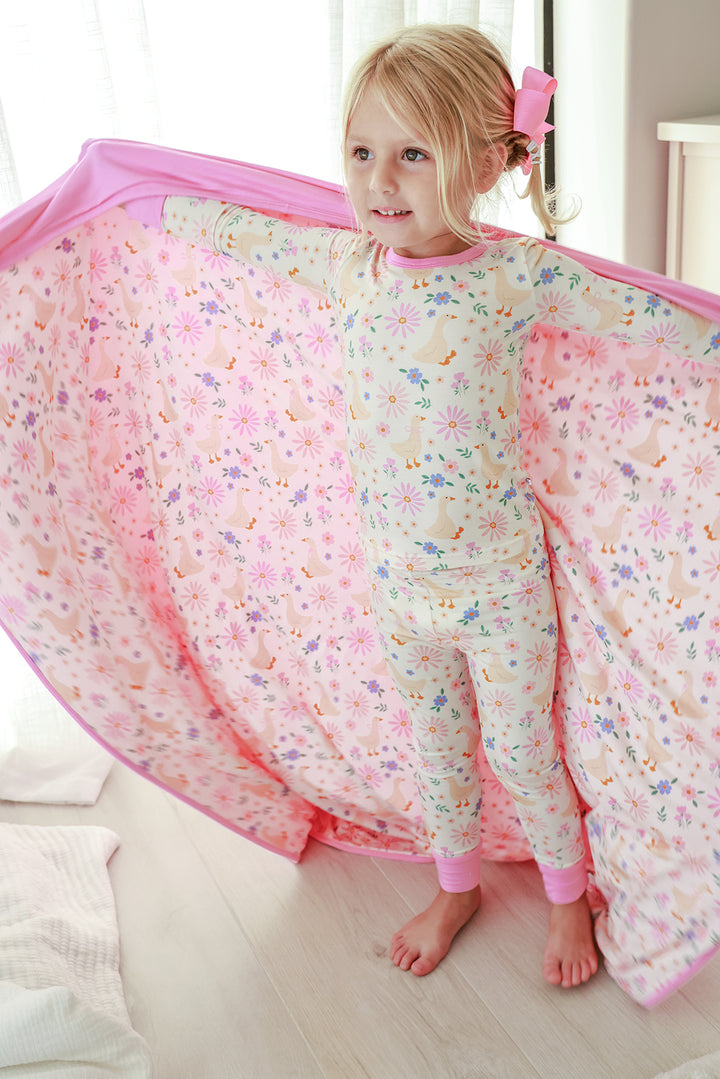 Young girl holding open the Double Sided Bamboo Blanket in the Lucy Goosy floral print showing pink solid reverse, wearing matching Lucy Goosy pajamas, lifestyle photo