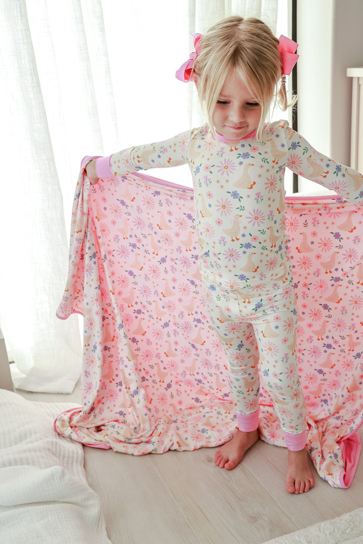 Young girl holding the Double Sided Bamboo Blanket in the Lucy Goosy floral print, wearing matching Lucy Goosy print pajamas, pink bow in hair, lifestyle photo