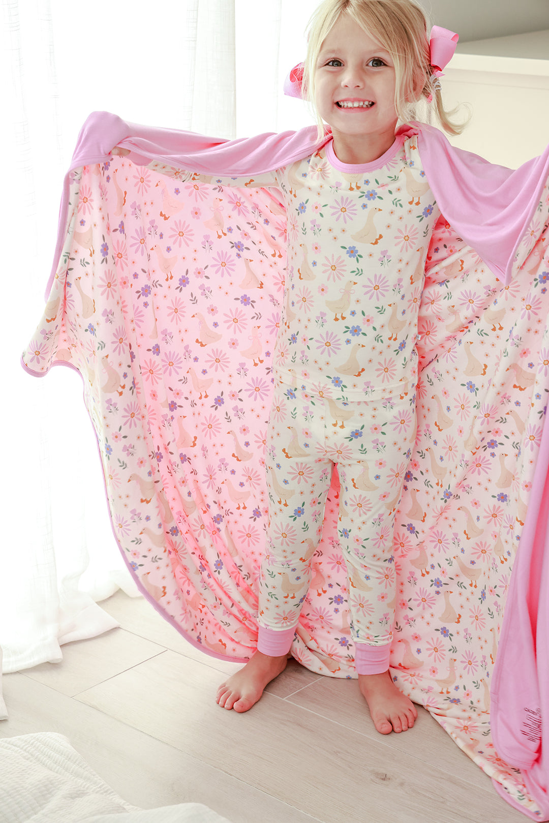 Young girl smiling and holding open the Double Sided Bamboo Blanket in the Lucy Goosy floral print showing the pink solid reverse, wearing matching pajamas, lifestyle photo