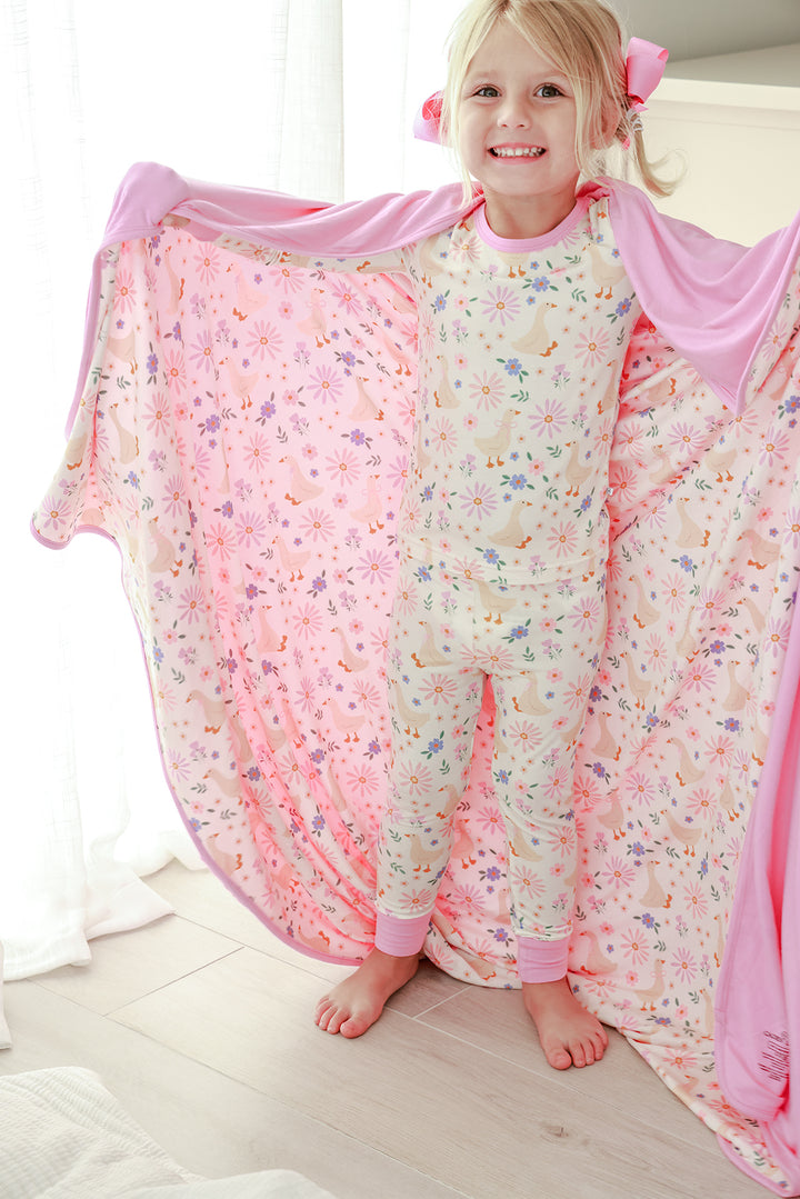 Young girl smiling and holding open the Double Sided Bamboo Blanket in the Lucy Goosy floral print showing the pink solid reverse, wearing matching pajamas, lifestyle photo