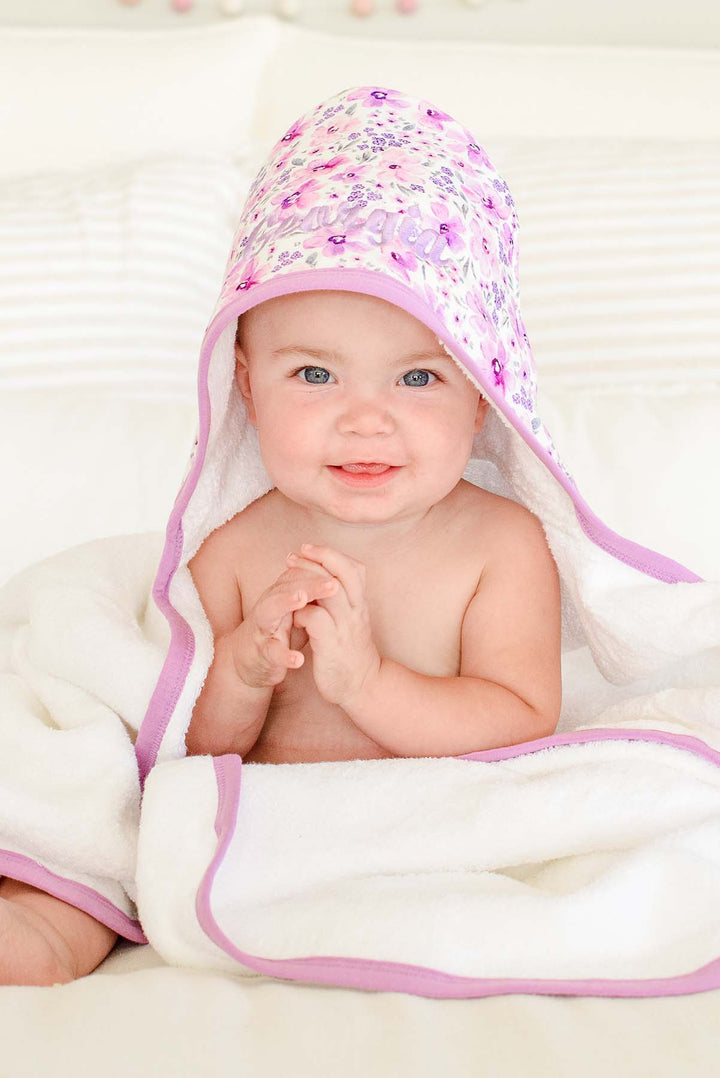 Smiling baby with hands clasped, wearing a Caden Lane white terry hooded towel in a Lilac Dreams print featuring pink watercolor blooms and grey leaves, reading "Georgia."