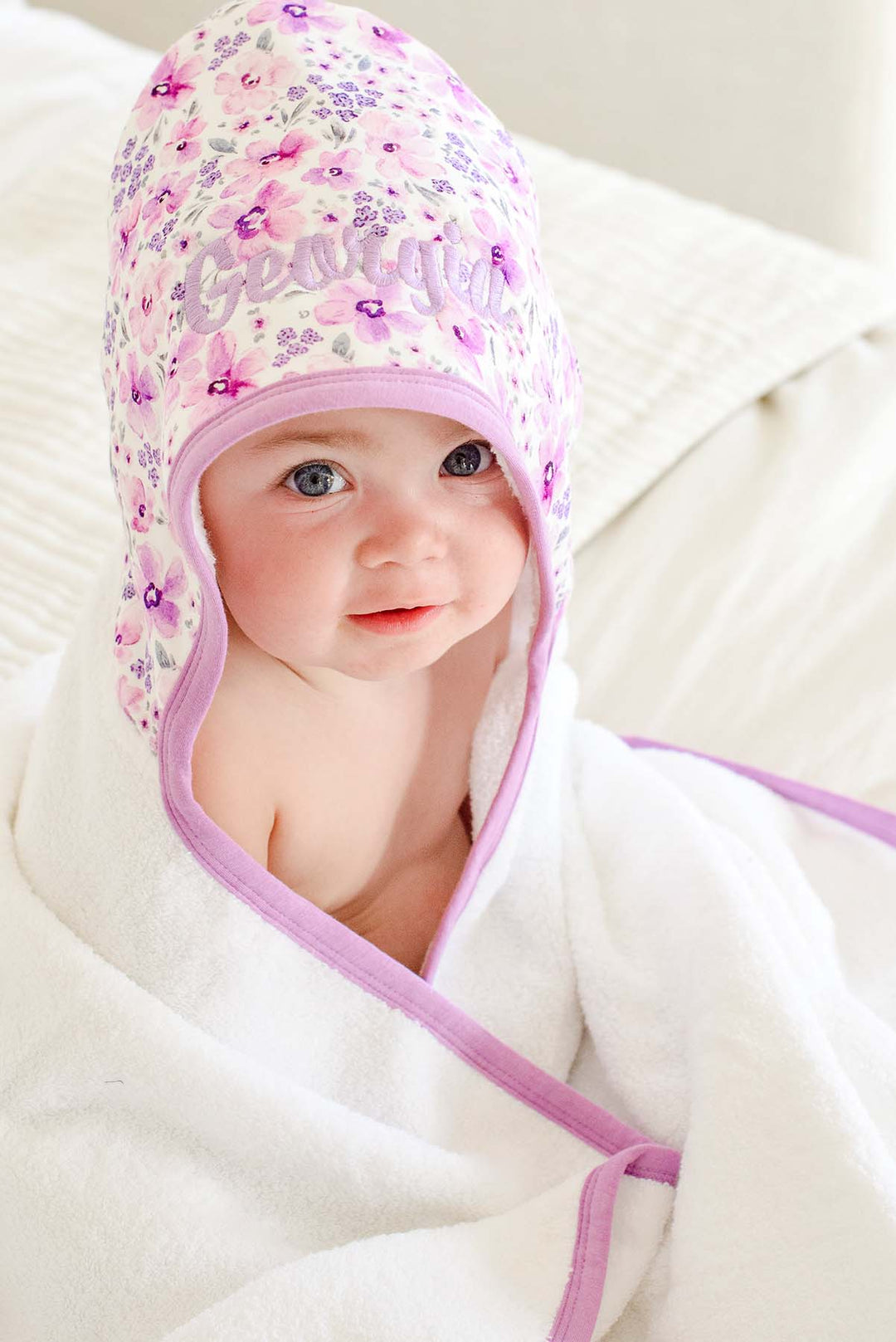 Angled shot of a baby gazing upward wearing a Caden Lane white terry hooded towel in a Lilac Dreams print featuring lavender florals, with lilac embroidery reading "Georgia."