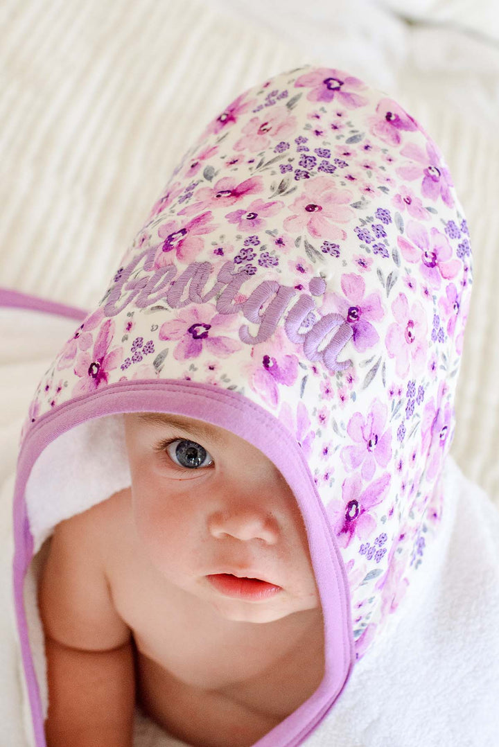 Close-up of a baby peeking out from under the hood of a Caden Lane white terry hooded towel in a Lilac Dreams print featuring purple watercolor flowers, reading "Georgia."