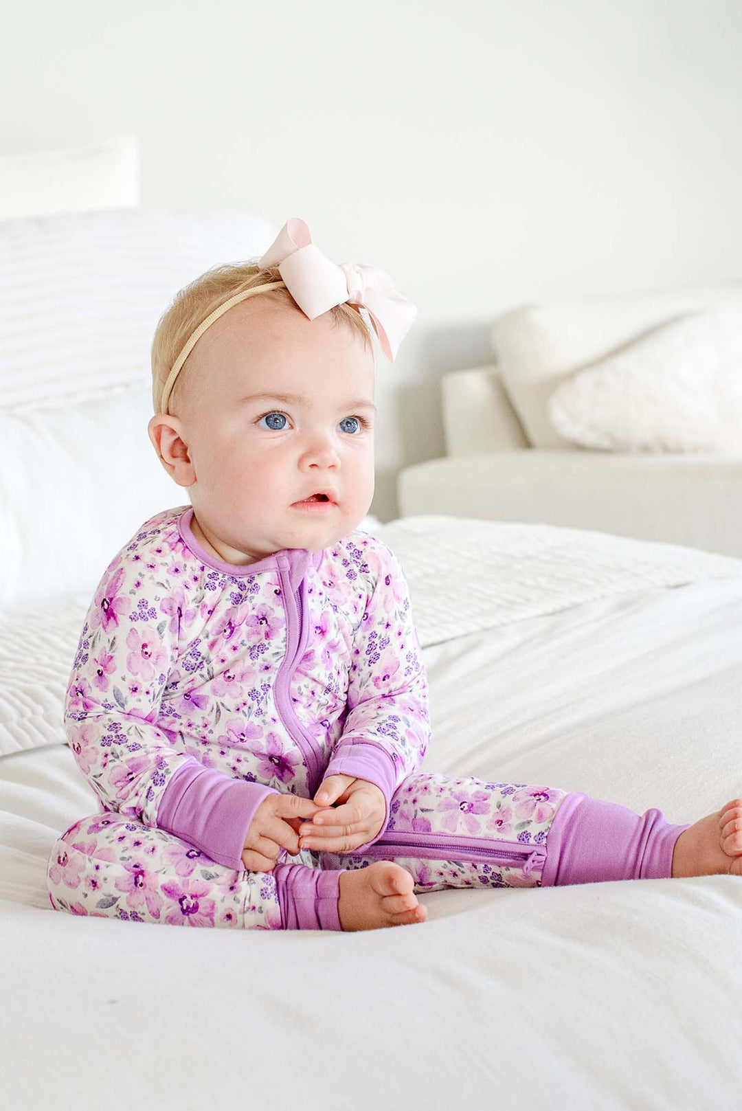 Seated baby with a pale pink bow gazing off camera, wearing a Caden Lane white knit zip romper with lilac cuffs in a Lilac Dreams print featuring purple watercolor flowers.