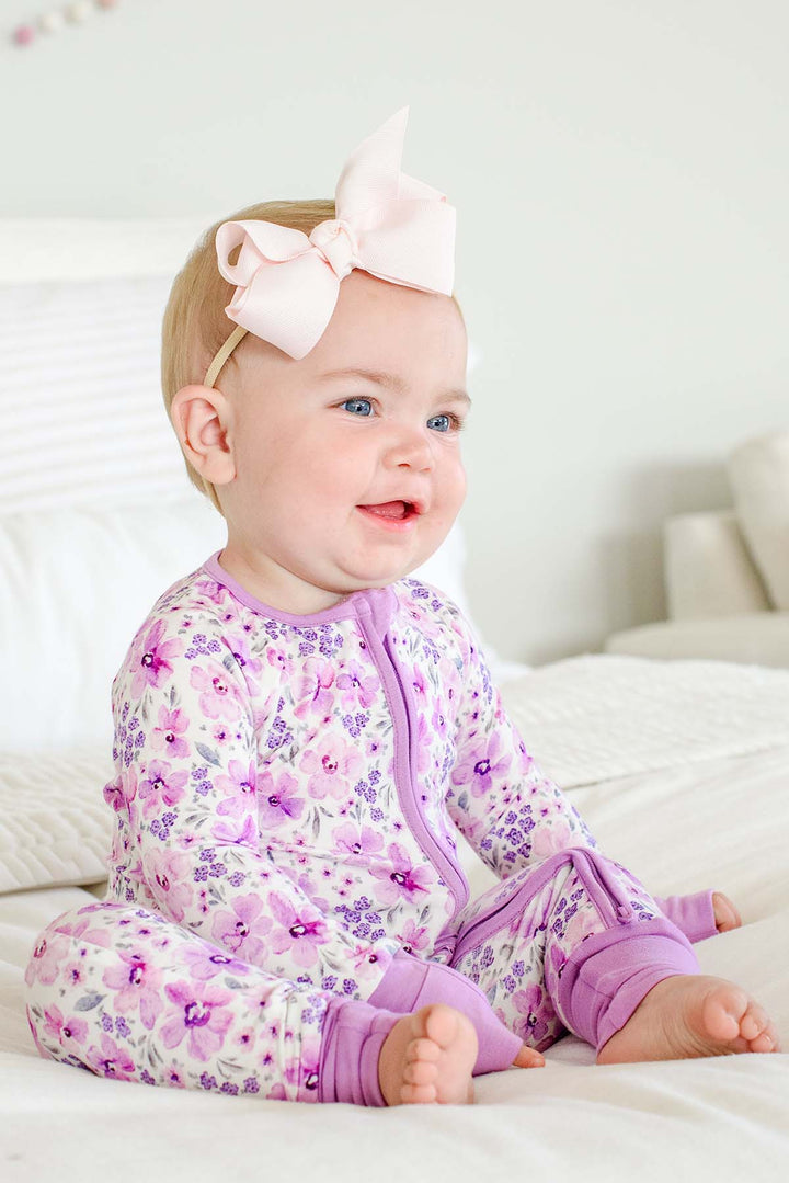 Smiling baby seated on a bed, wearing a Caden Lane white knit zip romper with lilac cuffs in a Lilac Dreams print featuring lavender florals and grey leaves.