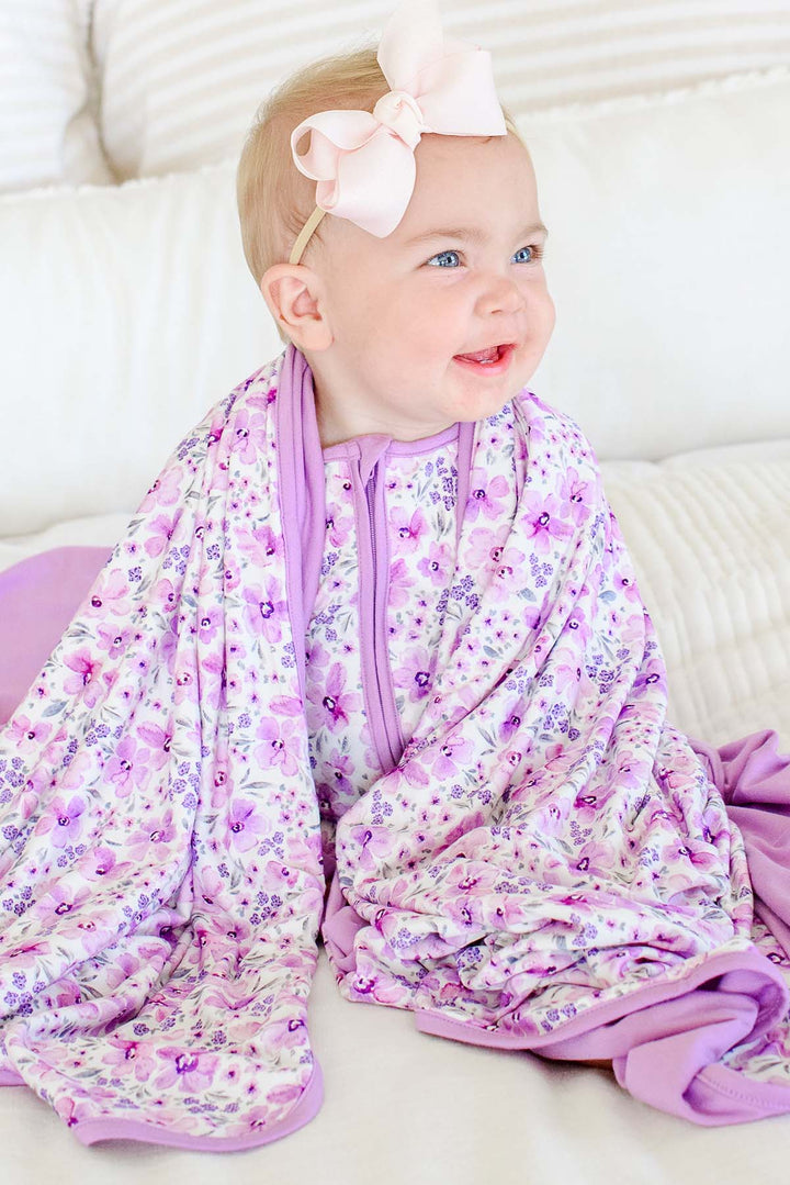 A smiling baby in a pink bow headband wrapped in a Caden Lane cream knit blanket in a Lilac Dreams print featuring watercolor flowers, paired with a matching romper.