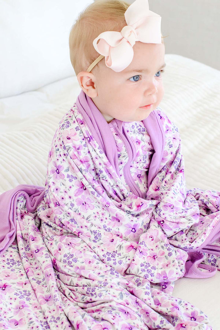 A baby gazing to the side, draped in a Caden Lane cream knit blanket in a Lilac Dreams print featuring watercolor flowers, paired with a pink hair bow.
