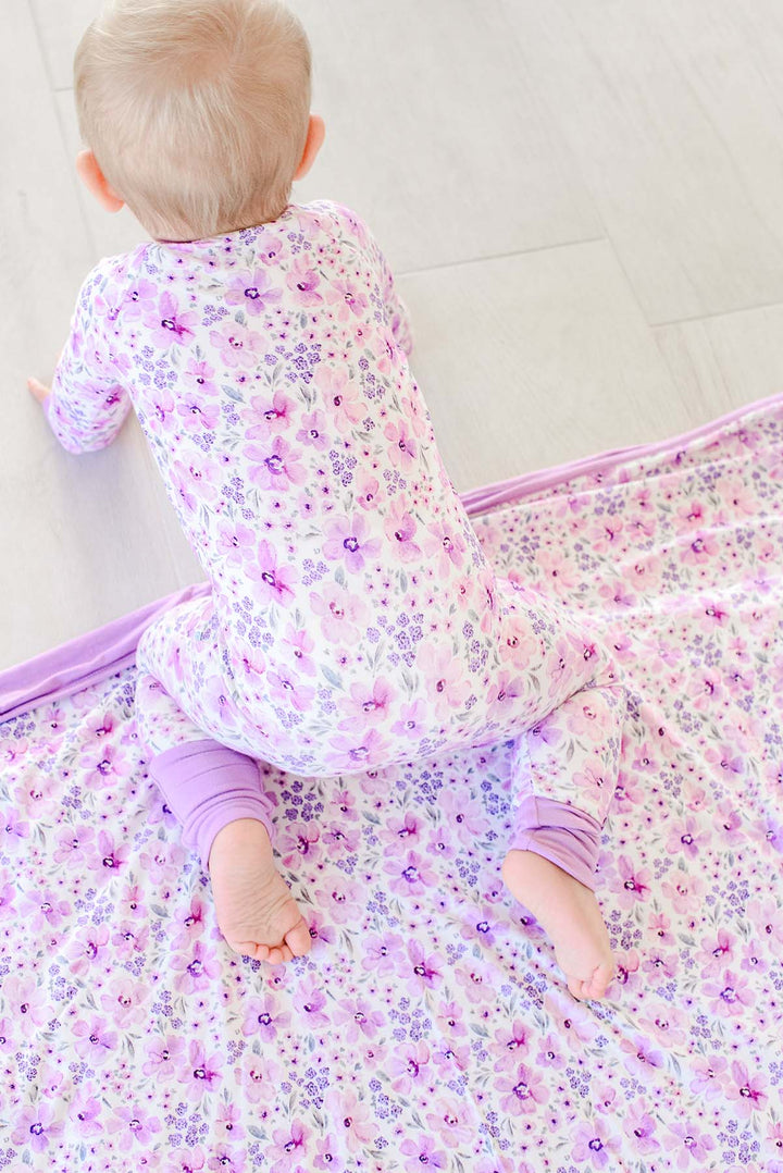 Back view of a crawling baby on a Caden Lane cream knit blanket spread across the floor in a Lilac Dreams print, paired with a matching romper.